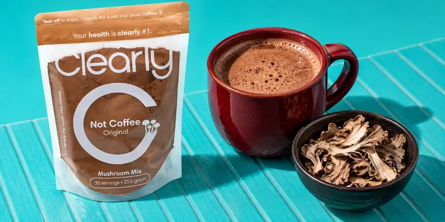 Busta di Clearly Not Coffee con una tazza di bevanda al cacao e una ciotola di fette di maitake essiccato su fondo turchese